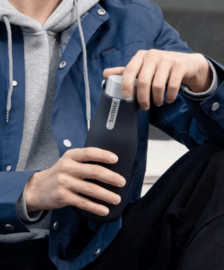 Ampolla purifica l'aigua, blau marí a les mans d’un home amb dessuadora gris, amb una jaqueta blava a sobre i pantalons negres.